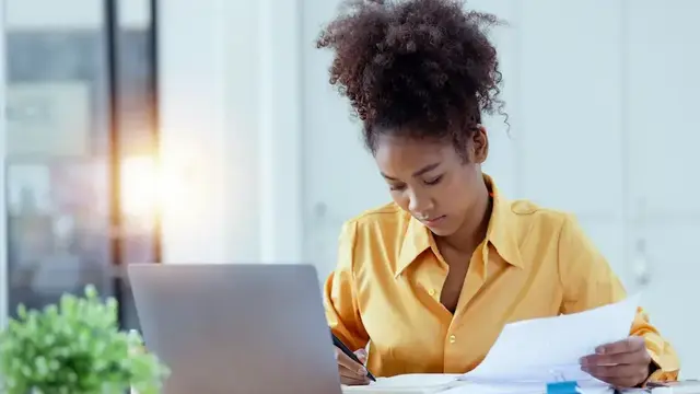 Office professional reviewing documents and taking notes while working on a laptop.