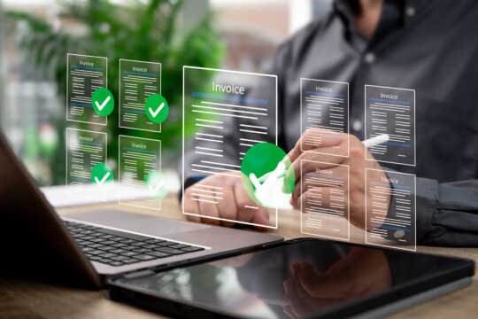 Digital invoice documents with checkmarks displayed over a laptop and tablet on a desk.