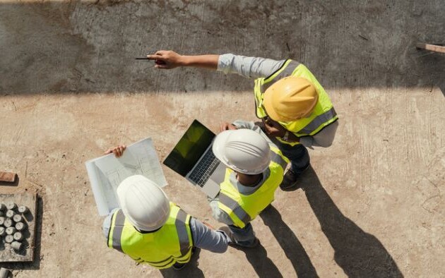 Overhead view of people wearing safety vests and hard hats reviewing paper plans and a laptop at a construction site.