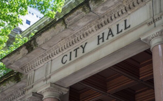 Stone entrance of a city hall building with columns and the words ‘City Hall’ carved above the doorway.