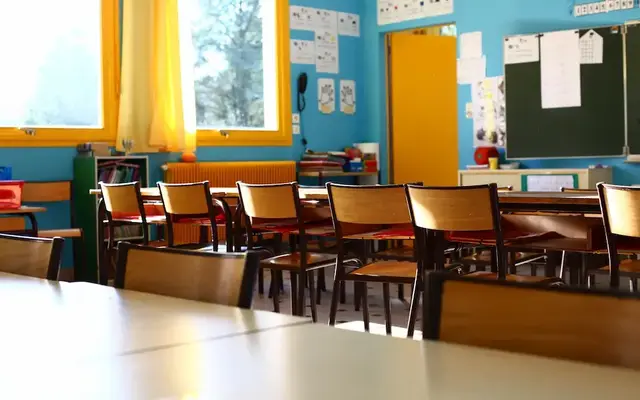 Bright, empty classroom with rows of wooden desks and chairs, colorful blue and yellow walls, large windows, and a blackboard at the front of the room.