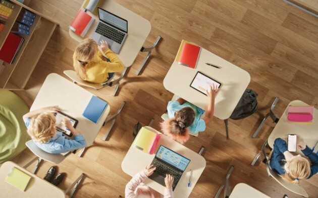 Overhead view of students seated at individual desks using tablets and laptops in a classroom.