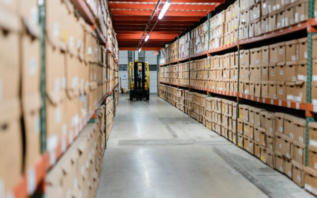 Long warehouse aisle with shelving filled with labeled storage boxes and a forklift in the distance.