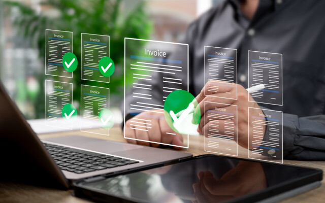 Digital invoice documents with checkmarks displayed over a laptop and tablet on a desk.