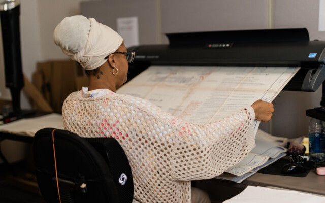 Person operating a large-format scanner with oversized paper documents in a production workspace.