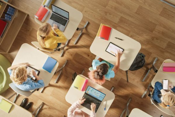 Overhead view of students seated at individual desks using tablets and laptops in a classroom.
