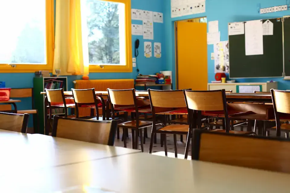 Bright, empty classroom with rows of wooden desks and chairs, colorful blue and yellow walls, large windows, and a blackboard at the front of the room.