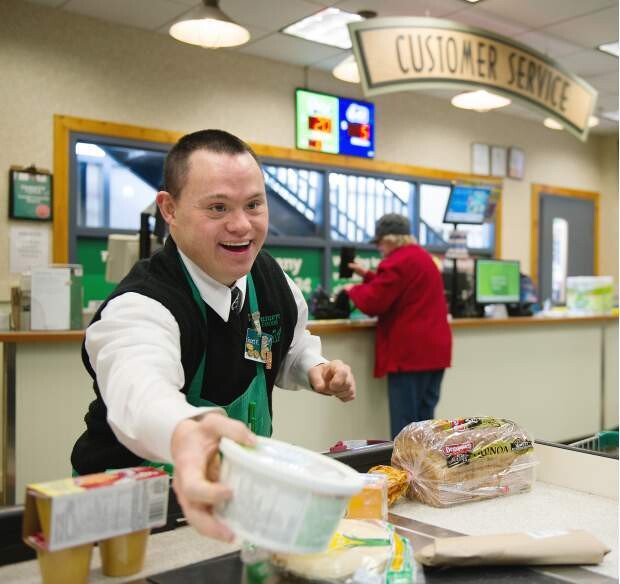 A smiling young man with a developmental disability is providing great customer service as he bags groceries at a supermarket