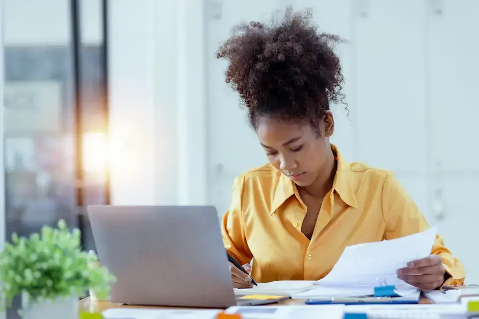 Office professional reviewing documents and taking notes while working on a laptop.