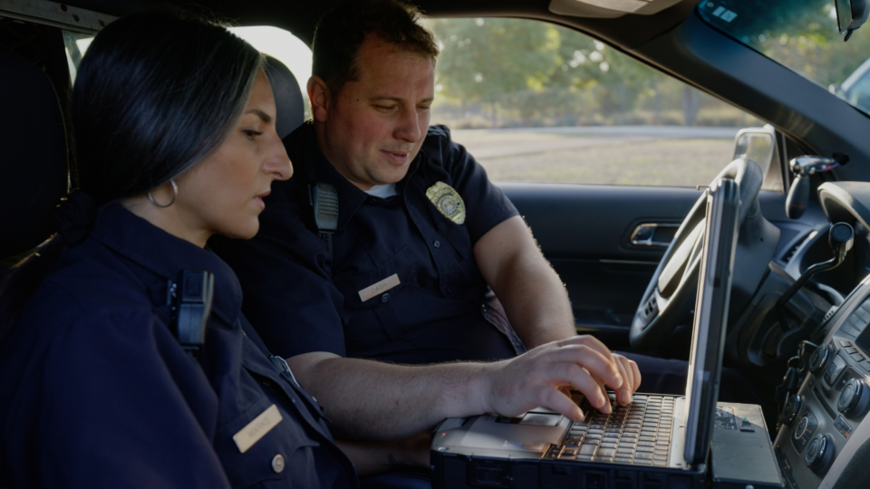 A male and a female police officer in their squad car looking at digital records on a laptop monitor.