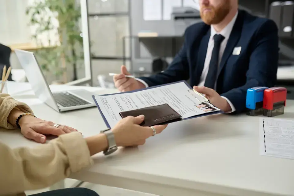 Two people in a government office are seated at a desk reviewing printed documents with a laptop and office stamps nearby.
