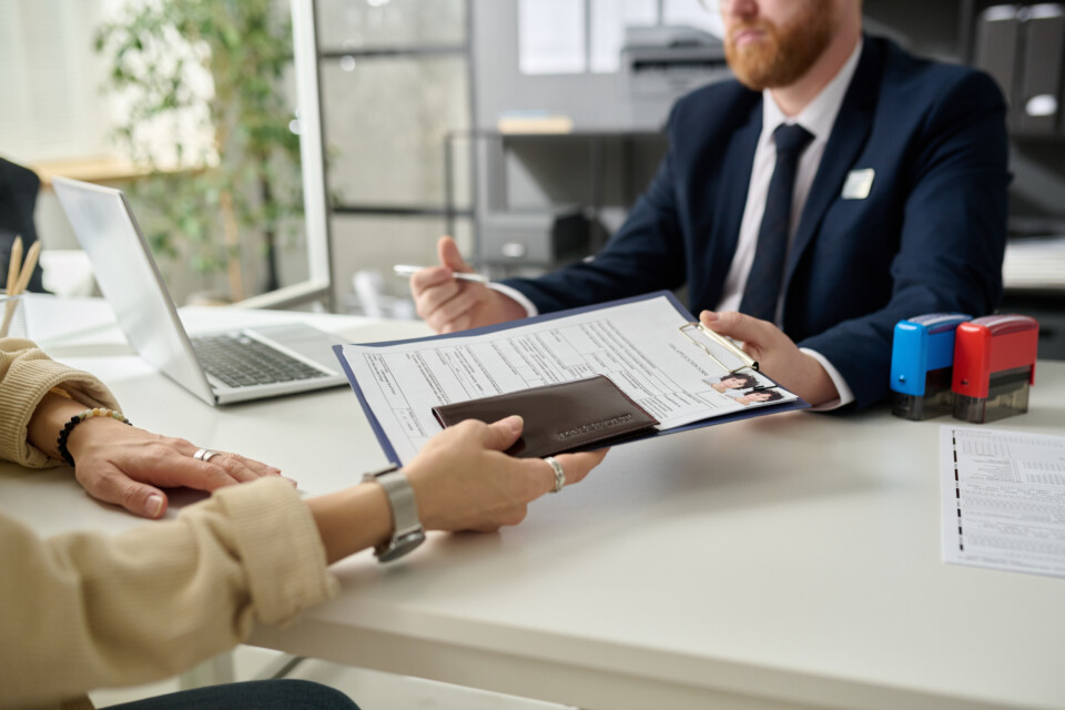 Two people seated at a desk reviewing printed documents with a laptop and office stamps nearby.