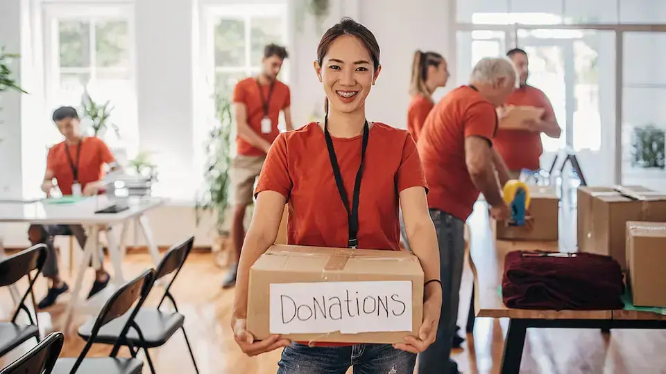 Nonprofit volunteer holding a box labeled “Donations” while other volunteers organize supplies in the background.