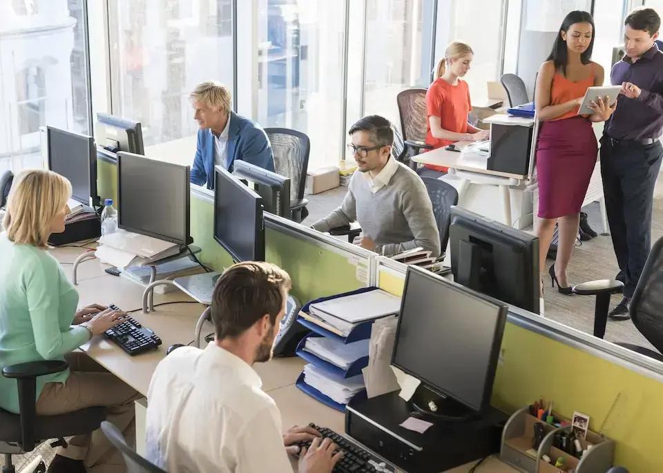 Employees working and collaborating in a modern open office with computers and shared workspaces.