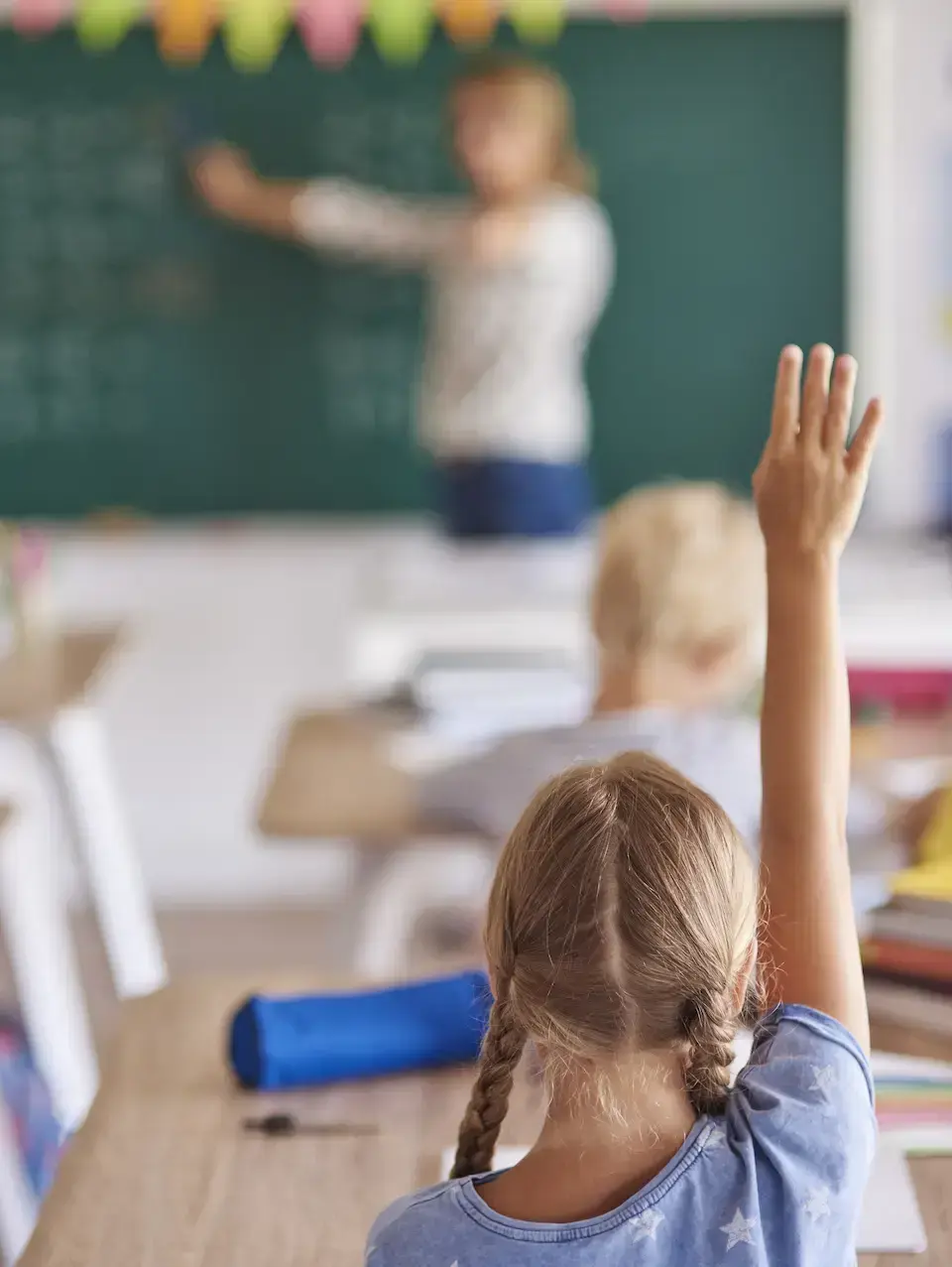 Student seated at a desk raising a hand in a classroom while an instructor writes on a chalkboard.